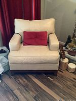Front view of cream leather chair with red microfiber pillow on the seat, on wood flooring with burgundy curtains in background.