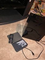 Kindle e-reader in case on carpet near black subwoofer and Sony Blu-ray player on shelf.