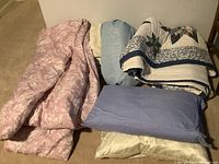 Photo of pink patterned comforter, blue textured coverlet, folded quilt with blue floral and geometric design, blue pillowcase, and beige satin pillowcase arranged on floor.