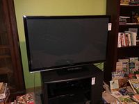 Front angled view of the Panasonic TV on a stand with remote and power cord visible, set against a green wall with books and board games nearby.