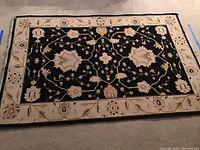 Full view of the rectangular rug showing black center with beige floral vine design and beige border with floral patterns.