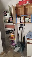 Wooden shelving unit with various gardening supplies and tools organized in and around it, including grout, joint compound, tomato cages, flower pots, flower bulbs, weed control sprayer, potting soil, and snow shovels.