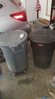 Photo showing two Rubbermaid trash cans with lids and wheels, one gray (Brute) and one black (Roughneck), in a garage next to a car.