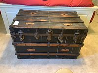 Front view of flat topped wood trunk with visible wear, metal braces, and tarnished brass hardware.