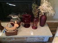 Wide view of ceramic urn vase, ruby glass vases, purple vase, and woven baskets on a stone fireplace hearth.