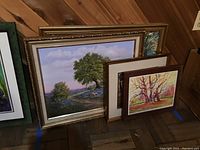 Four framed artworks arranged on floor against wood panel wall. One painting shows a large green tree with landscape and blue sky.