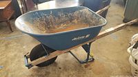 Side angled view of wheelbarrow showing the blue steel tray, wooden handles, and black pneumatic tire. Visible dirt and scratches on tray exterior.