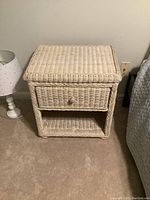 Front view of white wicker side table showing drawer with round knob and lower open shelf, placed next to a white lamp and bed.