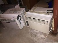 Two white window air conditioner units placed on a concrete basement floor showing the side and front of units.
