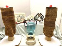 Full view of two Modern Cat cardboard scratching posts each topped with a red ball, pet waterer with blue reservoir and white base, two cat-themed oven pads, and two decorative cat plates visible in background.