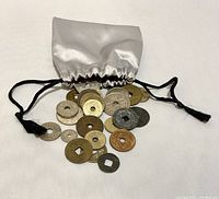 Photo of a silver satin drawstring sack with coins spilling out, showing various coins mostly round with holes in the center, different metal colors and sizes.