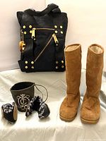 Photo of Juicy Couture black and gold seatbelt bag, honey brown suede boots, and black travel slippers on white background