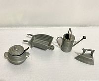 Four vintage metal miniature items arranged on white surface showing iron, pot with lid and spoon, watering can, and wheelbarrow.