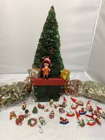 Full view of the miniature vintage Christmas tree with the assortment of small holiday ornaments arranged in front, and two bags of red and gold garland positioned on either side.
