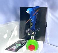 Photo of the front of the beer tap handle featuring the blue shark holding a pool cue standing on an 8-ball on a green base, with original packaging and brochure behind it.