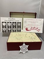 Box open displaying 44 white metal snowflake ornaments arranged in slots, with reference guide and poem sheets upright behind the box, and one ornament hanging from the box edge.