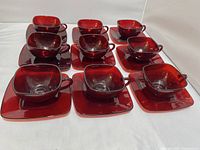 Full set of ruby red glass Anchor-Hocking Charm tea cups and saucers laid out on a white surface, showing 9 cups and 11 saucers.