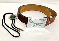 Photo showing the bolo tie and the tooled leather belt coiled, adjacent with the detailed buckle and bolo clasp visible.