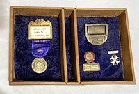 Photo showing all four vintage badges arranged inside a wooden box with blue velvet lining, separated into two compartments by a removable wooden slat. Visible are name badges and pins of various sizes and designs.