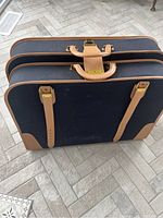 Full exterior view of navy blue canvas suitcase with tan leather trim, two leather straps with buckles, double handles folded down, set on tile floor.
