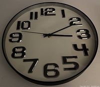 Photo showing front view of round wall clock with black frame, white face, and bold silver and black numbers 1-12. The hour, minute, and second hands are visible.