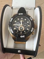 Front view of Invicta watch in holder, showing black dial, bezel with white numerals, gold-tone case, and black silicon strap.