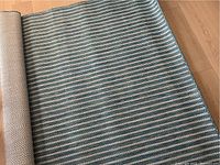 Top view of the rolled edge of a turquoise and white striped indoor/outdoor rug on a wooden floor showing weave and pattern details.
