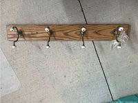 Full view of the solid oak coat rack showing the four brass double hooks with white ceramic knobs, placed on a concrete floor for clarity of the rack.