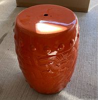 Orange ceramic stool or side table with textured decorative patterns and a drainage hole on top, photographed on concrete floor with natural lighting.