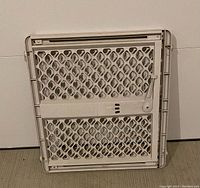 Folded white safety gate made of plastic and metal with diamond patterned grid and locking mechanism.