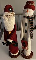 Photo of two Christmas decorative figures: Santa Claus and a snowman, both standing upright with festive holiday attire and holding small gift bags.
