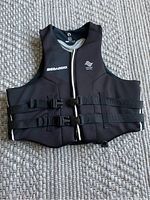 Black Sea Doo life vest showing front with zipper and two adjustable side buckles, laid flat on a textured surface.