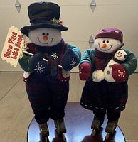 Two snowmen figurines on ice skates, one holding a wooden sign and the other holding a baby snowman, standing on a reflective surface with a garage door background.