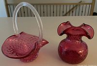 Photo of two vintage cranberry glass pieces on a light surface; one is a basket with a clear handle and the other a vase with a ruffled top.