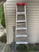 Front view of Werner 6 ft aluminum step ladder with five steps and red top, beside a black square nosed shovel