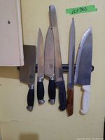 Six knives hanging on a wall, various blade types and handle colors (black, white, wood)