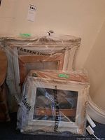 Two white ornate carved wood frames wrapped in plastic leaning against wall