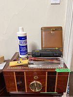 Full view of the wood inlaid cigar humidor with brass lock and hygrometer on front, bottle of Brigham humidor solution, two black cigar trimmers, two clear cigar tubes with cigars, and box of matches on top of the humidor.