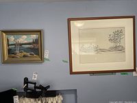 Pair of framed artworks on wall, showing landscape oil painting and signed print.