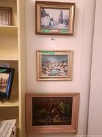 Three framed paintings on wall showing village scenes and still life. Each frame is different style and color.