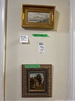 Two framed paintings hanging on a wall, the top one a watercolour of a Scottish mountain and loch landscape in a gold frame, the bottom one an oil painting of a cottage scene in a carved wooden frame.