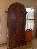 Full view of tall wooden armoire with rounded top, two large doors, and two lower drawers, positioned next to a window with an orange suitcase nearby.