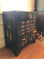 Front-left angled view of the black lacquered chest showing painted Asian figures and floral patterns, with metal lock on top.