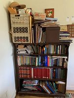 Bookshelf with multiple shelves packed with various books including novels, history, religious and reference books. Some file crates and miscellaneous items on top shelf.