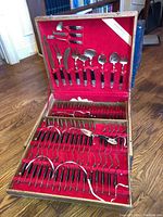 Full view of open wooden case showing cutlery pieces arranged on red lining including knives, forks, spoons, serving utensils.