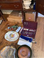 Photo showing full assortment of lot items on wooden floor including cutlery set in wooden case, ceramic platter, wooden bowls, soup terrine, serving utensils, and packaged utensils.