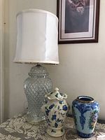 Glass ginger jar style lamp with white fabric shade and two ceramic urns on lace tablecloth