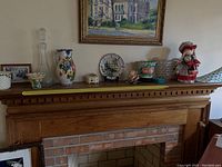 Wide shot of mantelpiece showing ceramic pitchers, flower pots, glass decanter, ceramic peacock figurine, and other pottery items arranged on top with framed pictures behind