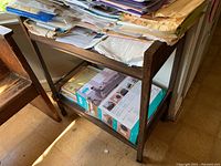 Wooden rolling cart seen from left side showing top shelf with various papers and files, and bottom shelf with boxed vacuum cleaner beneath.