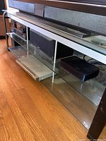 BellO TV stand showing clear glass top and two lower glass shelves supported by aluminum frame, with electronic components placed on shelves